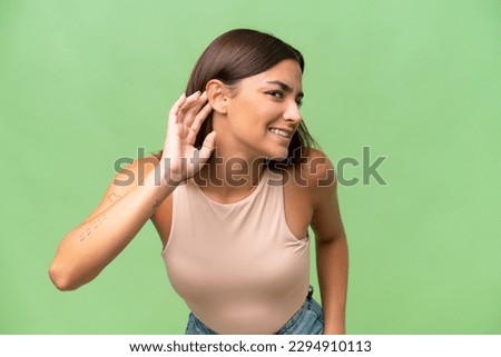 Young caucasian woman isolated on green chroma background listening to something by putting hand on the ear