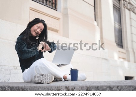 An Asian student makes a video call on a laptop computer, making a heart gesture with her hands. The young woman is sitting at the entrance of the university.