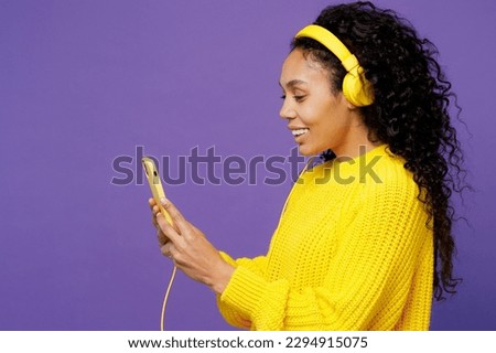 Side view young woman of African American ethnicity wear casual yellow sweater headphones listen to music use mobile cell phone isolated on plain purple background studio portrait. Lifestyle concept