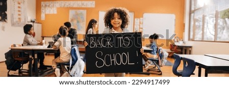 Happy boy holding a back to school sign on first day on his first day back at school. School kid looking at the camera and standing in front of his classroom, he is eager to start a new schooling year