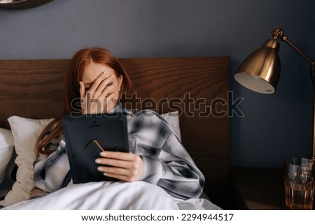 Portrait of grief-stricken young woman lying on bed covering face with hand and crying while holding picture frame, touching photograph with love. Concept of nostalgia, grief, longing and loneliness.