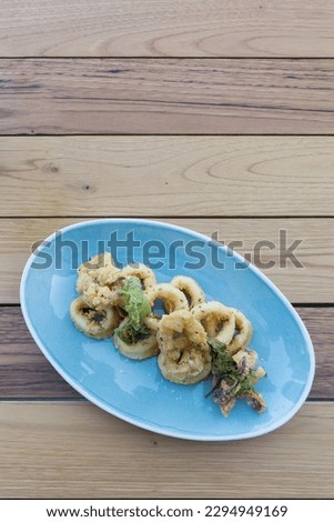 Isolated blue plate of breaded calamari rings, typically known by "calamares a la andaluza" in Spain, with edible green seaweeds on top. Picture of Mediterranean food on wooden background.