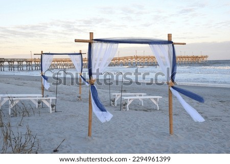 A couple wedding arbors and benches on the beach with a late after noon light and a fishing pier and ocean in the background.