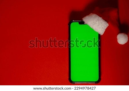 Mobile phone dressed in Santa-Claus red-white hat with chroma key screen against red background. Concept for Christmas or New Years holidays. Blank cell phone. Digital gadget wireless wishlist concept