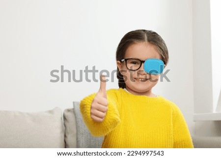 Happy girl with nozzle on glasses for treatment of strabismus showing thumbs up in room. Space for text Royalty-Free Stock Photo #2294997543
