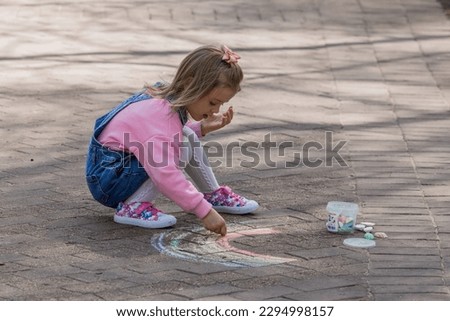 A happy girl in pink clothes draws a rainbow with chalk on the asphalt on the street. Portrait of a little girl drawing with rainbow chalk on a sunny summer day. Creative development of children.