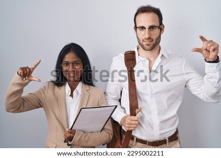 Interracial business couple wearing glasses smiling and confident gesturing with hand doing small size sign with fingers looking and the camera. measure concept. 