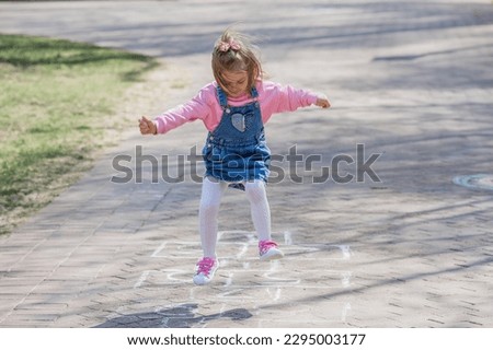 A happy girl in pink clothes draws classics with chalk on the asphalt on the street. Portrait of a little girl playing and jumping hopscotch on a sunny summer day. Creative development of children.