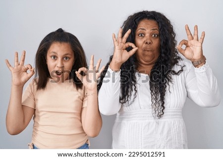 Mother and young daughter standing over white background looking surprised and shocked doing ok approval symbol with fingers. crazy expression 