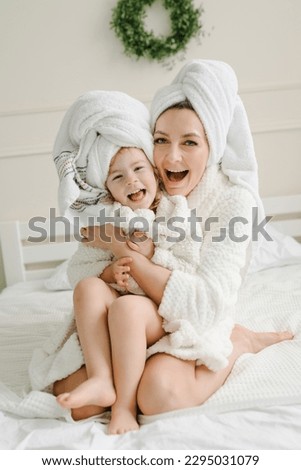 Young mother with cute little daughter in bathrobe, towels on heads sitting lovely on bed. Healthcare and family concept. Cheerful girls having beauty day at home. Happy mom hugging kid. Closeup.