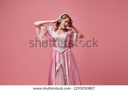 Modern princess. Shot of adorable blond princess, queen wearing fancy dress and listening music over pink studio background. Concept of gadgets, medieval, beauty, old-fashioned clothes, ad