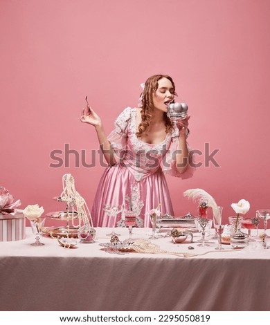 Celebrating life. Fairy tale blond princess wearing renaissance dress standing near the laid festive table with ice cream over pink studio background. Concept of medieval, beauty, party, baroque, ad