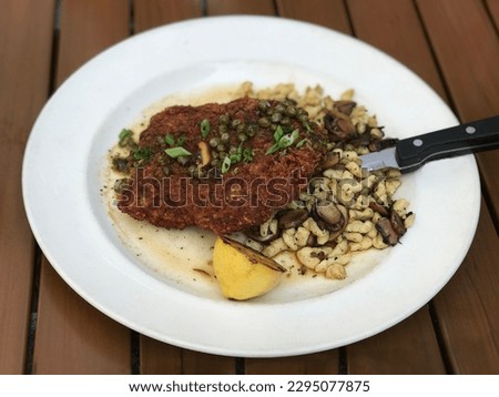 Fried chicken schnitzel with lemon and spatzle pasta with mushrooms is shown on a plate. Additionally, this updated version of the German classic is served with capers and green onions on the chicken.
