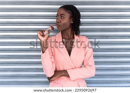 Young woman wearing a coral blazer dress posing in front of a silver roller shutter door while holding her sunglasses