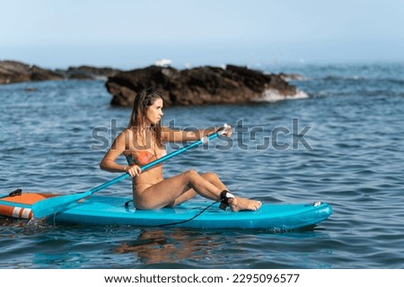 horizontal wide shot of an attractive, slim, tanned Caucasian brunette girl in a bikini, paddling her paddle surfboard in the sea water, with rocks in the background.