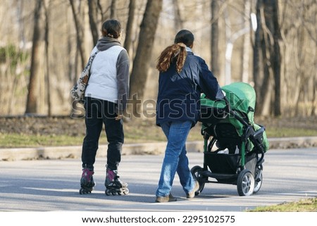 Mother with small child walks in the park. Her friend roller skating. Time for leisure and walking. Street photography. Backs, rear view