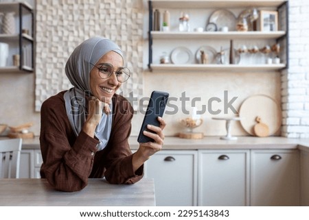 Close up photo. Smiling Muslim young woman in hijab standing in kitchen at home and using mobile phone, reading news, checking social media, watching videos, photos, searching for information.