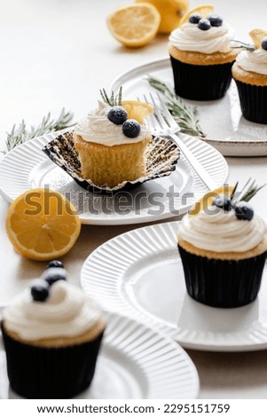 Lemon Cupcake with vanilla frosting and garnished with blueberries, lemon and rosemary. A collection of white a cream plates with rosemary sprigs, lemons and forks around the image.
