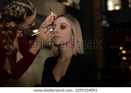 Makeup artist makes a professional evening make-up for a beautiful blonde woman. Creates a contour of the eyelids with brushes, paints with dark brown shadows. Concept. Beauty saloon.