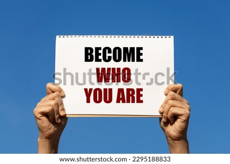 Become who you are text on notebook paper held by 2 hands with isolated blue sky background. This message can be used as business concept about becoming who you would like to be.