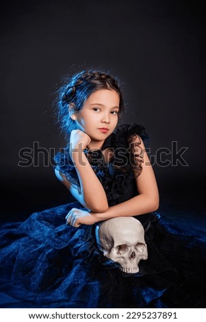 A little girl in a black dress with a pigtail hairstyle on her head poses sitting with a skull in her hands, isolated on a dark background with blue backlight.
