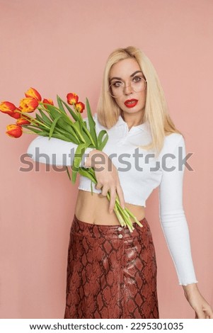 People emotions concept. Pretty adult woman stands indoor wears white shirt and brown snake skirt, round glasses isolated on pink background. Holding tulips in her hands. Springtime