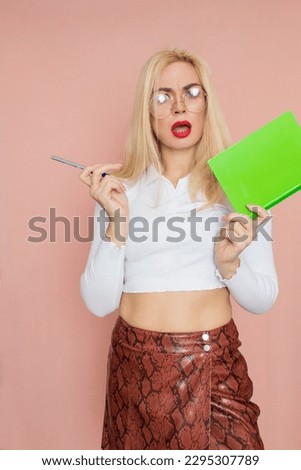 People emotions concept. Pretty adult woman stands indoor wears white shirt and brown snake skirt, round glasses isolated on pink background. Thinking about new ideas with book and pencil in her hands