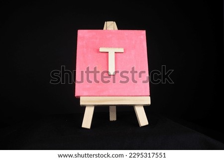 T wooden capital letter and pink blank painting canvas resting on a miniature artists easel isolated on a black background