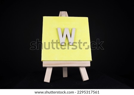 W wooden capital letter and yellow blank painting canvas resting on a miniature artists easel isolated on a black background