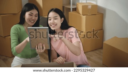 Asian pretty young woman with lovely friend enjoy to decoration in new home, sitting among many boxes and hold a photo frame in hand, They are discussing for idea to place it. move home concept