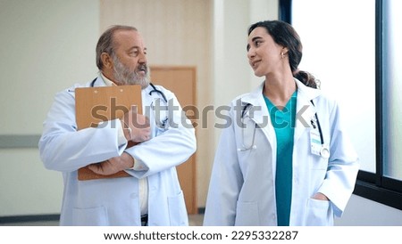 Two doctors wear white coats discussing diagnosis while walking through the hospital hallway. Young woman doctor talking to senior male doctor supervisor