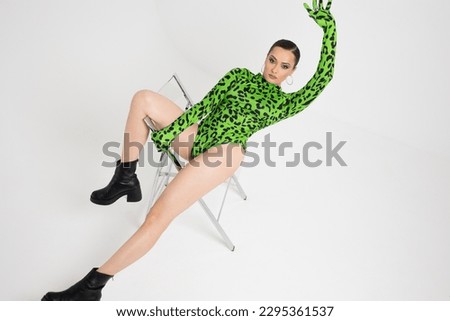Stylish bright dancer in a bright green leopard print bodysuit, posing on a chair, studio photo on a white background. Plastic body, pretentious pose