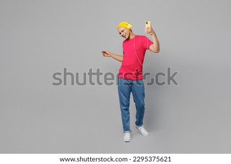 Full body young man of African American ethnicity 20s he wear pink t-shirt yellow hat headphones listen to music from own playlist use hold mobile cell phone dance isolated on plain grey background