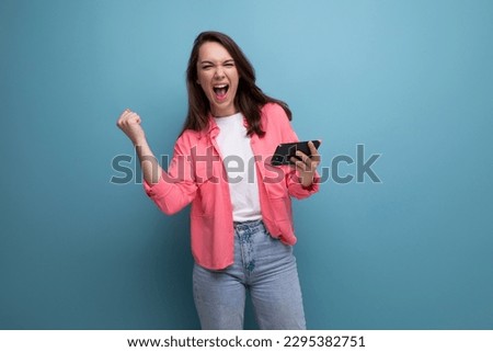 happy lucky brunette woman in informal look with smartphone on studio background Royalty-Free Stock Photo #2295382751
