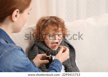 Mother giving cough syrup to her son on sofa indoors Royalty-Free Stock Photo #2295411713