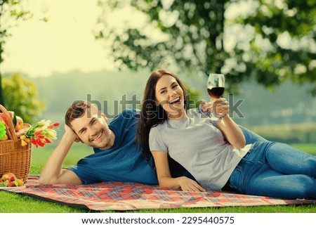 Bright sunny photo of happy excited young attractive couple in love, lying together on picnic blanket, with wine and basket. Cheerful man and woman outdoors.