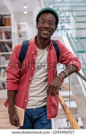Smiling contented african american student man in eyeglasses standing in university library look at camera. Portrait happy pleased black hipster guy with textbooks standing in college after lecture  Royalty-Free Stock Photo #2295496927
