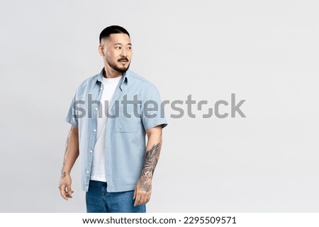 Portrait of attractive Asian man looking away at copy space, isolated on gray background. Young guy with stylish tattoo, casual clothes on the background of an empty wall, advertisement shopping