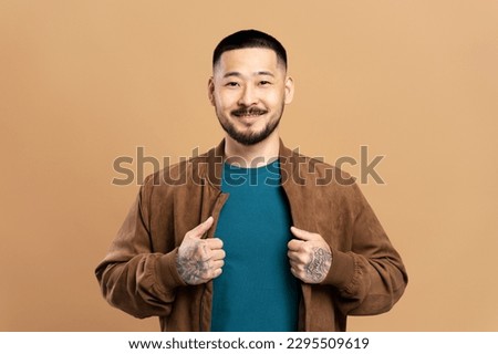 Portrait of smiling korean man in brown jacket standing isolated on beige background. Attractive businessman with stylish tattoos on arms looking at camera