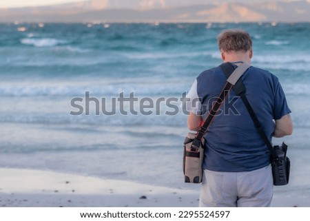 Mature man standing with his back to camera on beach shore, head lowered, two bags crossed on his back, sea and island at sunset in blurred background, El Tecolote beach, Baja California Sur, Mexico
