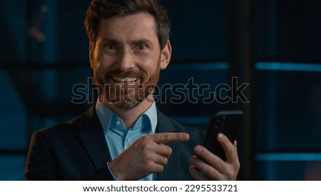 Caucasian bearded businessman happy 30s man worker entrepreneur winner stand in evening office hold cellphone click on mobile phone screen make online order recommend easy pay use gadget smartphone Royalty-Free Stock Photo #2295533721