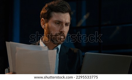 Caucasian 40s businessman employee auditor middle aged man sit in evening night office read business documents papers legal contracts pensive employee think check data in computer search information