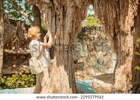 Elderly woman taking pictures on a mobile phone of a beautiful fountain and sculptures in a natural park.