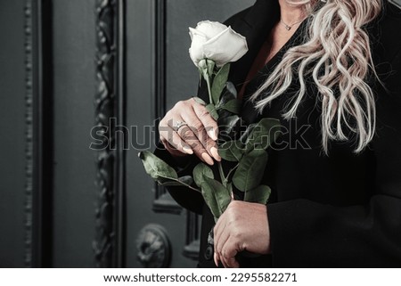 Close-up hands of pensive middle aged stylish woman holding white rose posing. Lonely sad alone mature lady standing near blackwall background. Loneliness emotion concept. Copy advertising text space