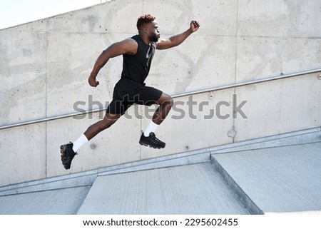 Confident strong fit sporty young black man running along urban wall. Motivated African ethnic guy jumping in air advertising sportswear, street workout training, pursuing goal concept. Side view Royalty-Free Stock Photo #2295602455