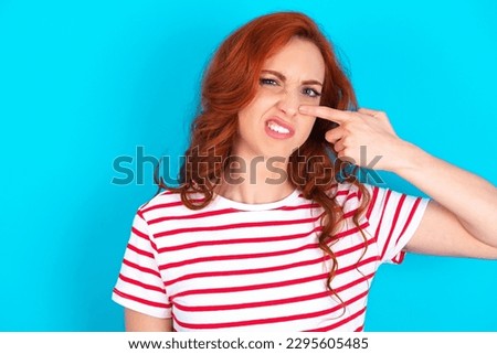 young redhead woman wearing striped T-shirt over blue background pointing unhappy to pimple on forehead, ugly infection of blackhead. Acne and skin problem
