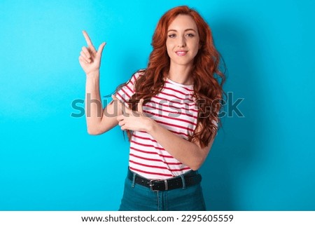 young redhead woman wearing striped T-shirt over blue background indicating finger empty space showing best low prices, looking at the camera