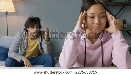 Annoyed caucasian husband sitting on couch and shouting at his asian wife, who is turned away from him, massaging her temples to calm down - domestic violence  Royalty-Free Stock Photo #2295638919