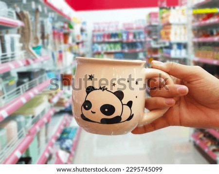 Holding a beige coffee mug with Panda drawing printed image and blur supermarket background.