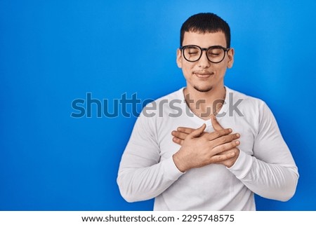 Young arab man wearing casual white shirt and glasses smiling with hands on chest with closed eyes and grateful gesture on face. health concept. 